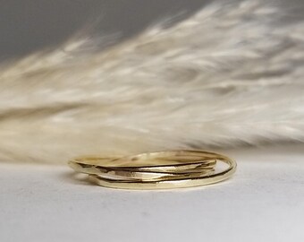Pode incluir: Uma pilha de três anéis de ouro delicados. Os anéis têm uma textura martelada e um design simples e elegante. Os anéis são empilhados sobre uma superfície branca, com um fundo desfocado de plantas secas e plumosas.