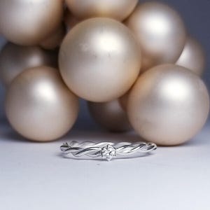 Könnte beinhalten: Ein silberner Ring mit geflochtenem Band und einem kleinen, klaren Edelstein in der Mitte. Der Ring ist vor einer Gruppe großer, goldfarbener Kugeln platziert, wodurch ein weicher, verschwommener Hintergrund entsteht.