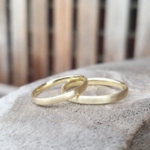 May include: Two gold wedding bands with a matte finish are displayed. The rings are placed on a light gray surface. The bands are simple, elegant, and classic, perfect for a wedding or commitment ceremony.