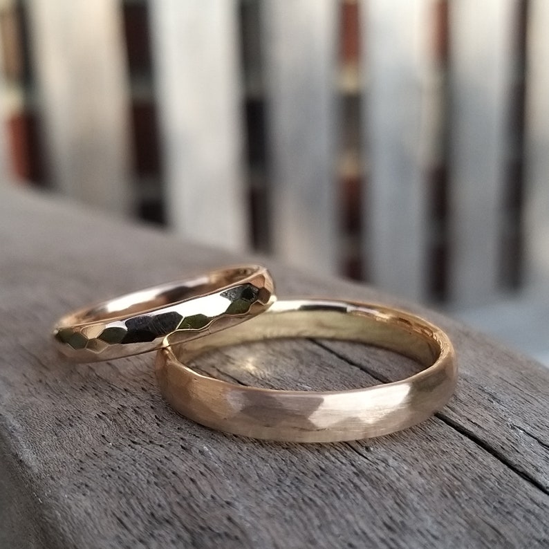 Puede incluir: Dos alianzas de oro, una con acabado martillado y la otra con acabado liso, descansando sobre una superficie de madera.
