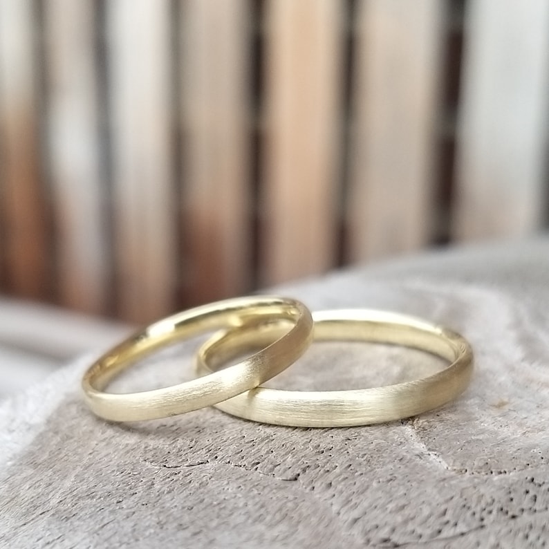 May include: Two gold wedding bands with a matte finish are displayed. The rings are slightly different in size, with one thinner than the other. The bands are set against a neutral, blurred background.