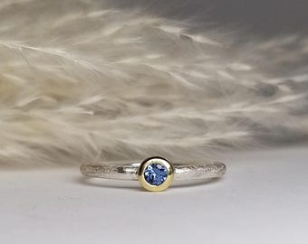 Op de afbeelding: Een zilveren ring met een getextureerde band en een blauwe edelsteen gezet in een gouden bezel.