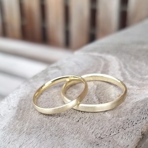 May include: Two gold wedding bands, one with a matte finish and the other with a polished finish, are interlocked on a weathered wooden surface. The rings are close together, suggesting a symbol of unity and commitment.