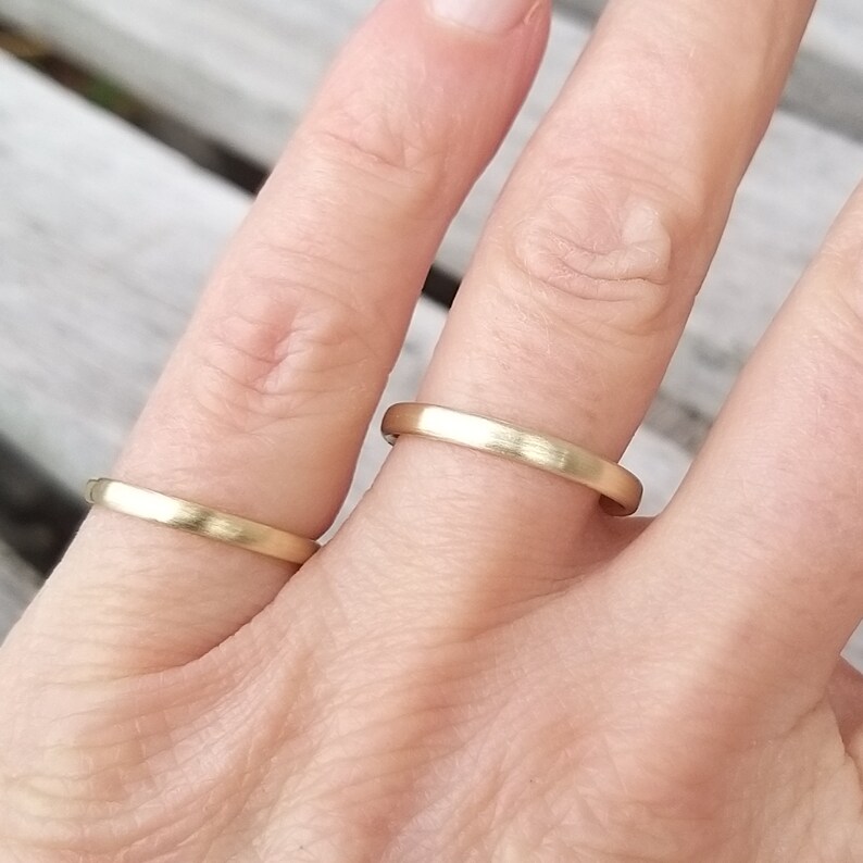 May include: Two gold wedding bands. The rings are a simple, classic design with a matte finish. One ring is on a finger, and the other is next to it. The rings are a warm, yellow gold color.