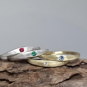 Könnte beinhalten: Drei einfache, gehämmerte Metallbänder mit jeweils einem einzelnen Edelstein. Die Bänder sind auf einem Stück Treibholz gestapelt. Das obere Band ist silber mit einem roten Edelstein, das mittlere Band ist silber mit einem grünen Edelstein und das untere Band ist gold mit einem blauen Edelstein.