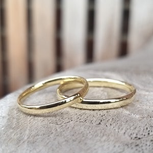 May include: Two gold wedding bands, one slightly overlapping the other. The rings have a polished finish and a simple, classic design. The bands are set against a soft, textured background.