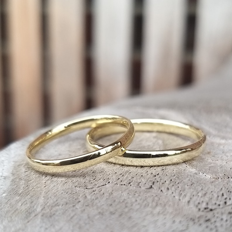 May include: Two gold wedding bands, one slightly overlapping the other. The rings have a polished finish and a simple, classic design. The bands are set against a soft, textured background.