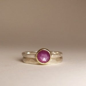 Könnte beinhalten: Ein Silber- und Goldring mit einem rosafarbenen Edelstein. Der Ring besteht aus zwei Bändern, einem silbernen und einem goldenen, wobei der Edelstein in das goldene Band gefasst ist.