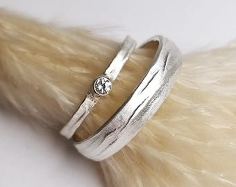 Pode incluir: Dois anéis de prata com um acabamento texturizado semelhante a casca de árvore. O anel mais fino apresenta uma pequena pedra preciosa redonda e clara engastada. O anel mais largo tem uma textura de superfície orgânica e irregular.