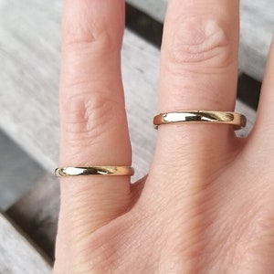 May include: Two gold wedding bands are displayed on fingers. The rings have a polished, shiny finish. The bands are simple, classic, and elegant, with a rounded profile. The rings are likely made of gold.