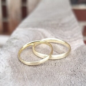 May include: Two gold wedding bands, one slightly overlapping the other, rest on a textured, light gray surface. The rings have a matte finish, and the soft lighting highlights their simple, elegant design. The bands are a symbol of commitment.