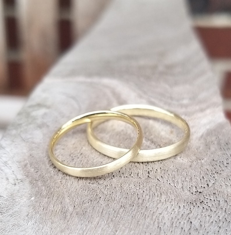 May include: Two gold wedding bands, one slightly overlapping the other, rest on a textured, light gray surface. The rings have a matte finish, and the soft lighting highlights their simple, elegant design. The bands are a symbol of commitment.