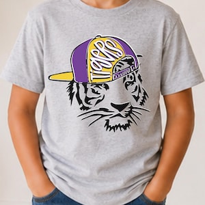 May include: A light gray short-sleeve t-shirt featuring a graphic of a tiger face wearing a purple and yellow baseball cap. The cap has the word Tigers printed in white script lettering across the front panel. The tiger is depicted in black.
