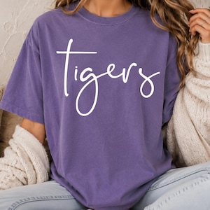 May include: A purple short-sleeved t-shirt featuring the word "tigers" written in white cursive script across the chest. The garment has a crew neckline and a relaxed fit, showcasing a solid color design with clear, legible white lettering.