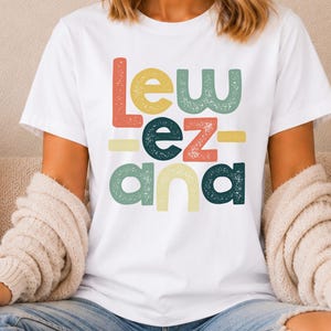 May include: A white short-sleeved t-shirt featuring the text "lew ez ana" printed in a stacked, multi-colored, distressed font. The letters are colored in shades of coral, sage green, dark teal, and pale yellow, arranged in three distinct rows.