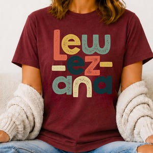 May include: A maroon short-sleeved t-shirt featuring a multi-colored graphic print with the text "Lew ez ana" in a distressed, retro-inspired font. The letters are arranged in three rows with varying colors including orange, yellow, teal, and red.