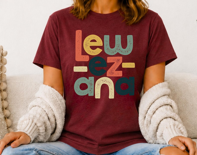 May include: A maroon short-sleeved t-shirt featuring a multi-colored graphic print with the text "Lew ez ana" in a distressed, retro-inspired font. The letters are arranged in three rows with varying colors including orange, yellow, teal, and red.