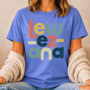 May include: A periwinkle blue short-sleeve t-shirt featuring the text "LEW EZ ANA" printed in a stacked, multi-colored, distressed font. The letters are colored in shades of coral, sage green, and dark teal, with small horizontal bars.