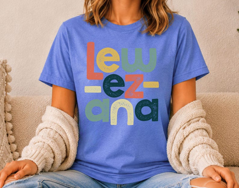 May include: A periwinkle blue short-sleeve t-shirt featuring the text "LEW EZ ANA" printed in a stacked, multi-colored, distressed font. The letters are colored in shades of coral, sage green, and dark teal, with small horizontal bars.