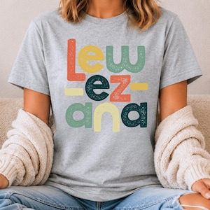 May include: A heather gray short-sleeve t-shirt featuring the text "LEW EZ ANA" printed in a stacked, multi-colored, distressed font. The letters are colored in shades of orange, yellow, green, and dark teal, arranged in a blocky, retro-inspired style.