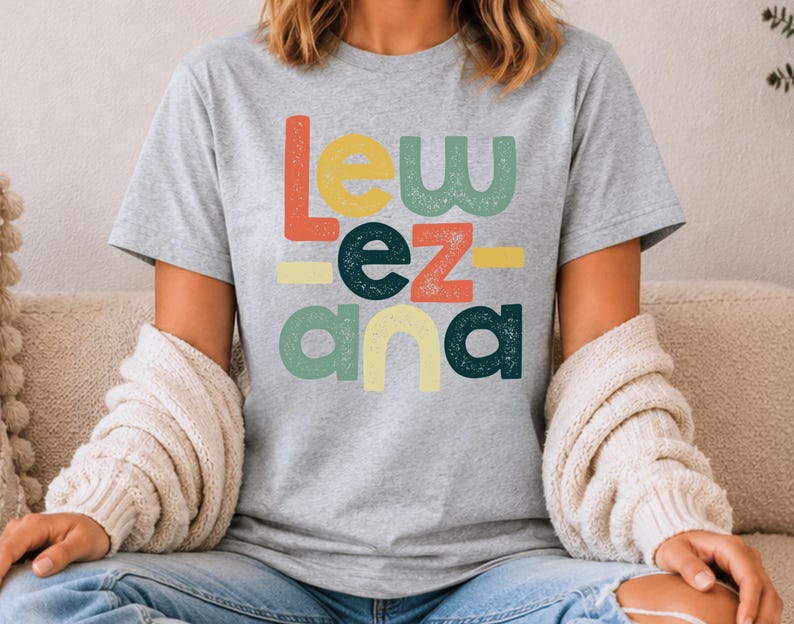 May include: A heather gray short-sleeve t-shirt featuring the text "LEW EZ ANA" printed in a stacked, multi-colored, distressed font. The letters are colored in shades of orange, yellow, green, and dark teal, arranged in a blocky, retro-inspired style.