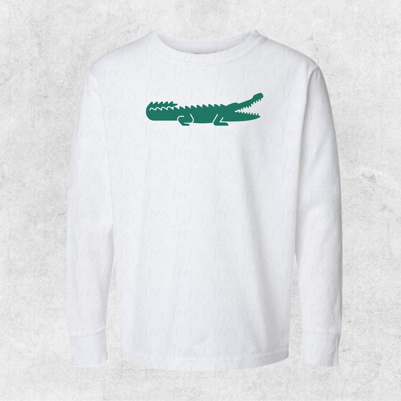 May include: White long-sleeved t-shirt with a green alligator logo on the front.