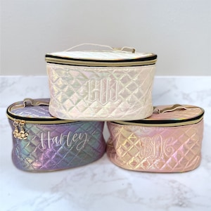 May include: Three quilted oval-shaped cosmetic bags with gold zippers and handles. The bags are in different colors: white, purple, and pink. Each bag has a different monogram embroidered on the front: "EJL", "Harley", and "BDC".