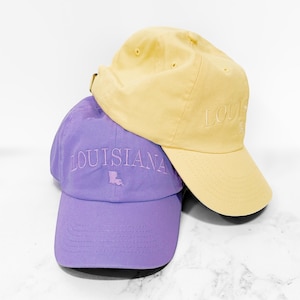 Pu&ograve; includere: Due cappellini da baseball, uno lavanda e uno giallo, sono impilati. Il cappellino lavanda ha la scritta "LOUISIANA" ricamata sul davanti, con un contorno dello stato. Il cappellino giallo ha la scritta "LOU" ricamata sul davanti. Entrambi i cappellini hanno una visiera curva.