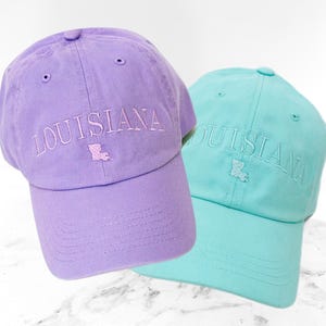 Pu&ograve; includere: Due cappellini da baseball, uno lavanda e uno verde acqua, sono esposti. La parola "LOUISIANA" &egrave; ricamata sulla parte anteriore di ogni cappellino, insieme a un contorno ricamato dello stato. I cappellini hanno una visiera curva e un bottone sulla parte superiore.