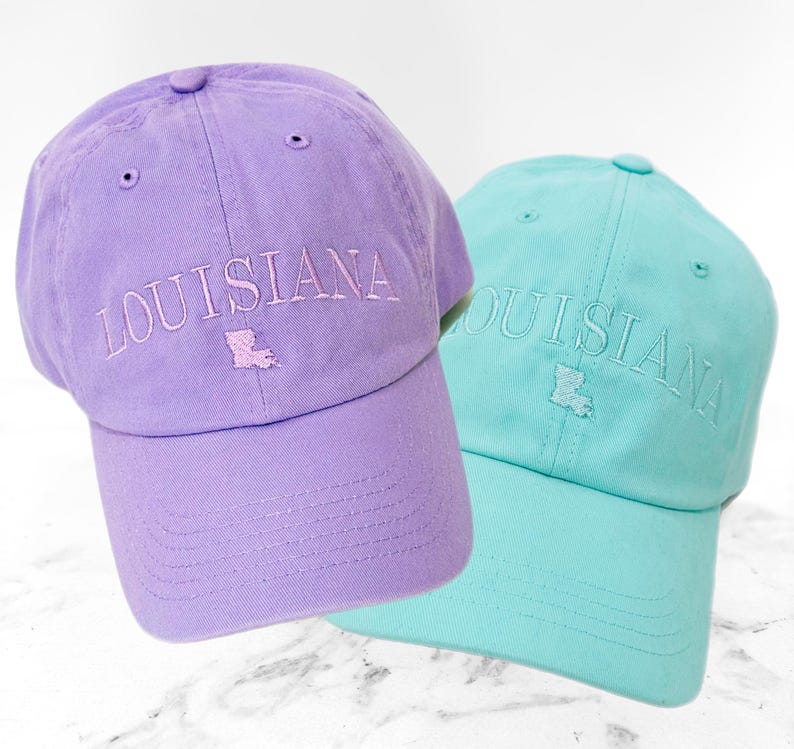 Pu&ograve; includere: Due cappellini da baseball, uno lavanda e uno verde acqua, sono esposti. La parola "LOUISIANA" &egrave; ricamata sulla parte anteriore di ogni cappellino, insieme a un contorno ricamato dello stato. I cappellini hanno una visiera curva e un bottone sulla parte superiore.