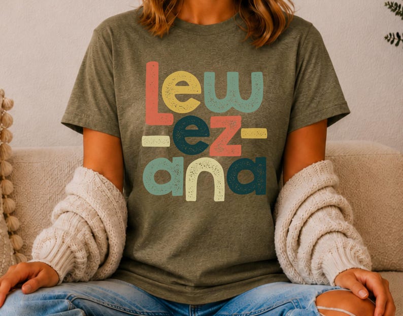 May include: An olive green short-sleeve t-shirt featuring the text "Lew ez ana" printed in a stacked, multi-colored, distressed font. The letters are colored in shades of coral, yellow, dark teal, and light teal, arranged in a blocky layout.