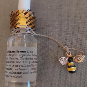 K&ouml;nnte beinhalten: Eine durchsichtige 10 ml Glas-Pipettenflasche mit Bienengift-Serum und einem goldenen Originalit&auml;tssiegel am Flaschenhals. Ein kleiner goldfarbener Bienenanh&auml;nger mit schwarz-gelben Streifen und klaren Strasssteinen an den Fl&uuml;geln h&auml;ngt an einer Schnur.