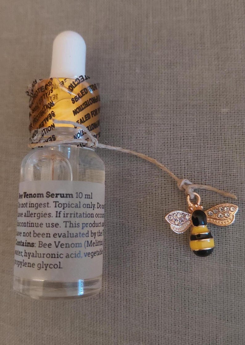 K&ouml;nnte beinhalten: Eine durchsichtige 10 ml Glas-Pipettenflasche mit Bienengift-Serum und einem goldenen Originalit&auml;tssiegel am Flaschenhals. Ein kleiner goldfarbener Bienenanh&auml;nger mit schwarz-gelben Streifen und klaren Strasssteinen an den Fl&uuml;geln h&auml;ngt an einer Schnur.