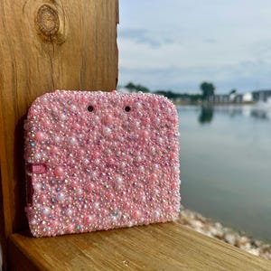 May include: A pink, bedazzled case, covered in small pink and iridescent beads. The case has a rectangular shape with rounded corners and a small pink clasp. The background shows a wooden surface and a body of water.