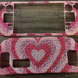 May include: A two-piece handheld gaming console shell set covered in small pink, magenta, and iridescent rhinestones. The front piece features a large heart pattern, while the back piece includes cutouts for buttons, joysticks, and vents.