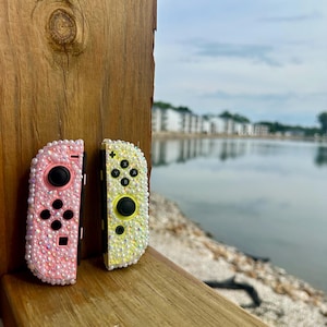 Op de afbeelding: Twee met juwelen versierde gamecontrollers, één roze en één geel, worden weergegeven op een houten oppervlak. De roze controller heeft een zwarte joystick en knoppen, terwijl de gele controller zwarte knoppen en een gele joystick heeft. De controllers zijn ingelegd met glinsterende edelstenen.