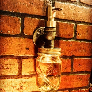 May include: A mason jar with a metal lid and the word "Mason" etched on the front is attached to a brass faucet. The faucet is mounted on a brick wall. The jar is filled with a clear liquid.