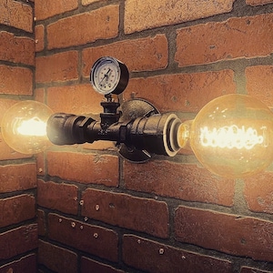 May include: A black metal pipe wall sconce with two light bulbs and a round gauge. The sconce is mounted on a brick wall.