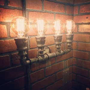 May include: Four Edison light bulbs mounted on a black metal pipe fixture against a brick wall. The light bulbs are turned on and glowing.