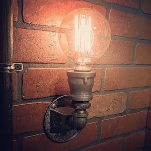 May include: A black metal pipe light fixture with a clear glass bulb mounted on a brick wall. The light is turned on and the bulb is glowing.