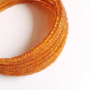 May include: A close-up of a beaded bracelet made of many small, orange beads. The bracelet is coiled into a circle and is lying on a white surface.