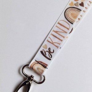 Puede incluir: Cordón blanco con la frase "Be Kind" en escritura a mano, junto con diseños de arcoíris y corazones marrones. Un cierre de metal plateado está unido al extremo del cordón.