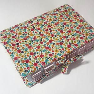 May include: A floral-patterned journal with a colorful design featuring red, yellow, and blue flowers on a white background. The journal has a matching fabric ribbon closure and tabbed pages.
