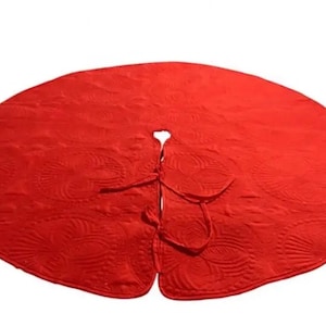 May include: A circular red fabric tree skirt featuring a quilted floral pattern across the surface. The item has a central opening and a slit extending to the edge, secured by two matching red fabric ties at the top for closure around a base.