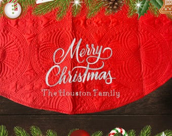 May include: A red Christmas tree skirt with the words "Merry Christmas" embroidered in silver. The skirt has a quilted pattern and the family name "The Houston Family" is also embroidered. Festive decorations surround the skirt.