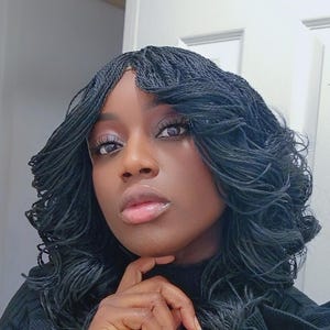 May include: A woman with dark brown skin is wearing a black mock neck shirt and has long, black micro braids styled in loose curls. She is looking at the camera with a serious expression.