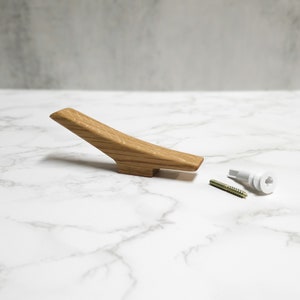 May include: A light wood coat hook with a curved design. The hook is shown on a white marble surface with a white plastic anchor and a gold screw.
