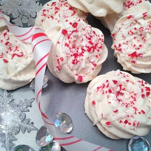 May include: White meringue cookies topped with white chocolate and crushed candy canes. The cookies are arranged on a silver tray with a red and white striped ribbon.