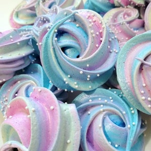 May include: A close-up of several pastel-colored meringues. The meringues are swirled with shades of blue, pink, and purple, and are decorated with small white sprinkles. The treats are arranged in a pile, showcasing their delicate texture.
