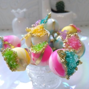 May include: A collection of colorful cake pops decorated with edible glitter and gold accents. The cake pops are arranged in a glass vase on a white surface.